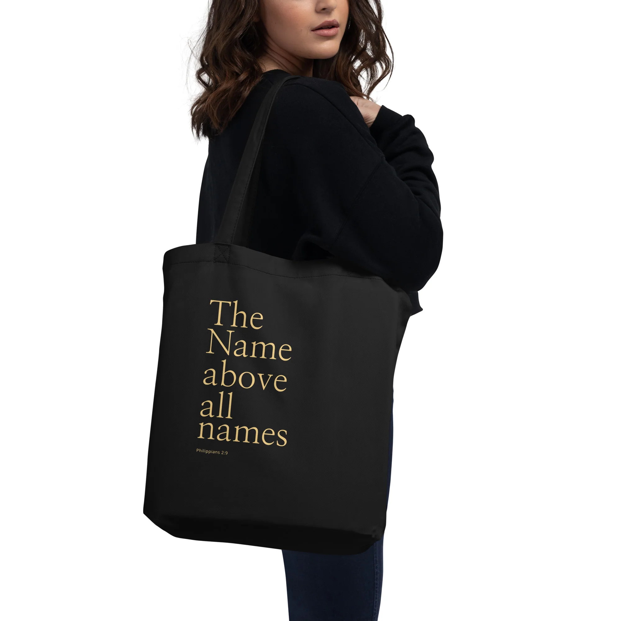 Model carrying the black King of Kings tote bag printed with gold “The Name above all names” typography